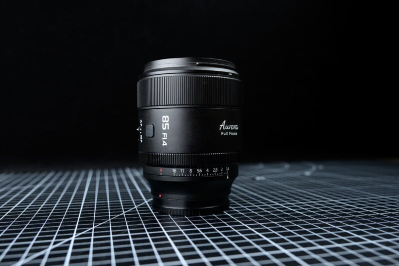 A black camera lens labeled "85 F1.4 Auroras Full Frame" stands upright on a black and white grid surface against a dark background.