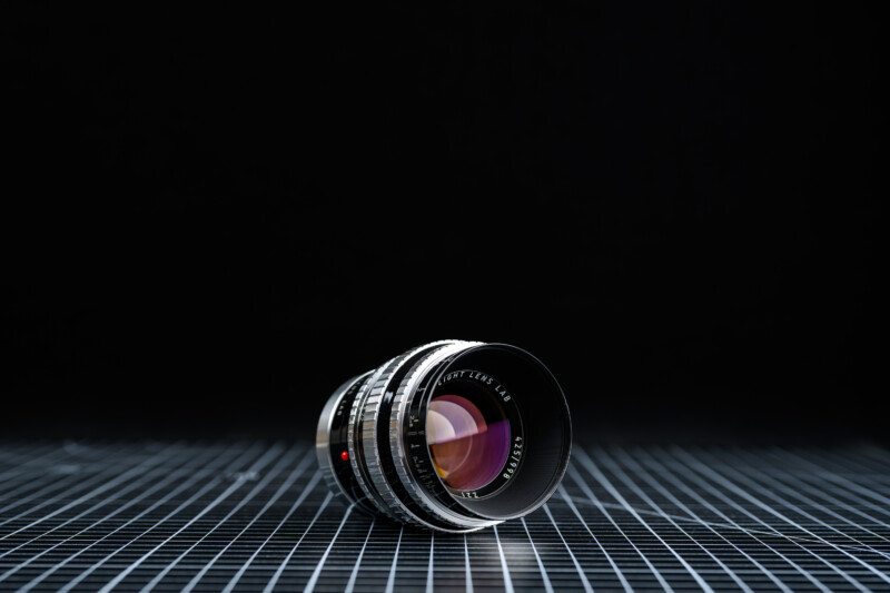 A camera lens lies on its side on a black grid-patterned surface, with a dark, out-of-focus background. The lens is detailed and reflective, with visible glass elements and engraved markings.