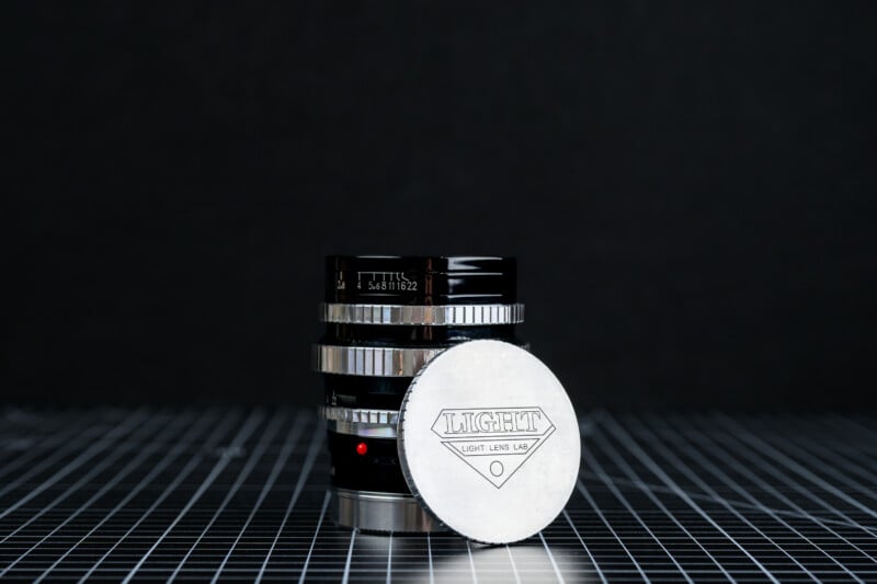 A camera lens with metallic rings stands on a grid-patterned surface. In front of it, a silver lens cap with the word "LIGHT LENS LAB" engraved is propped upright. The background is dark and plain.