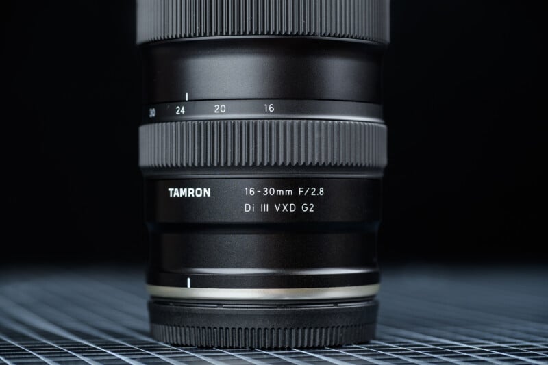 A close-up of a Tamron 16-30mm F/2.8 Di III VXD G2 camera lens, showing focus and zoom rings, with a black background and a grid pattern beneath the lens.