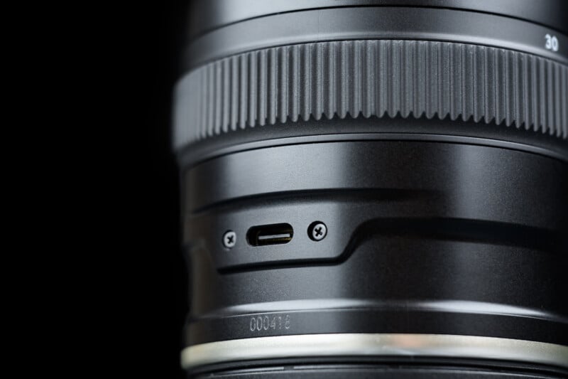 Close-up of a camera lens showing a textured focus ring, a micro USB port with two screws, and a section of the lens barrel with engraved numbers, all set against a black background.