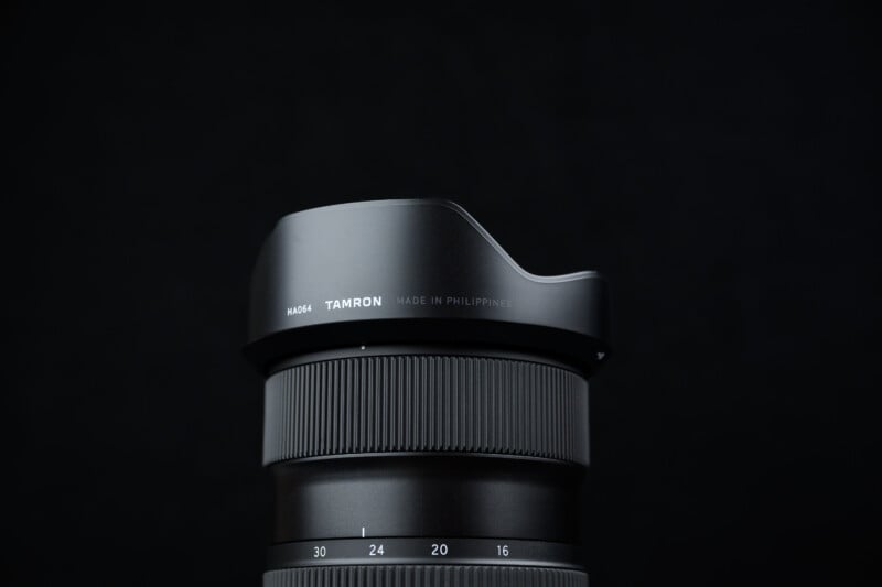 A close-up of a Tamron camera lens with a petal-shaped lens hood, set against a black background. The text "Tamron" and "Made in Philippines" are visible on the lens hood.