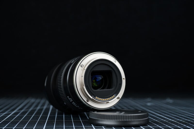 A camera lens lies on its side on a checkered surface, with its rear element facing forward and a lens cap placed beside it, set against a dark background.