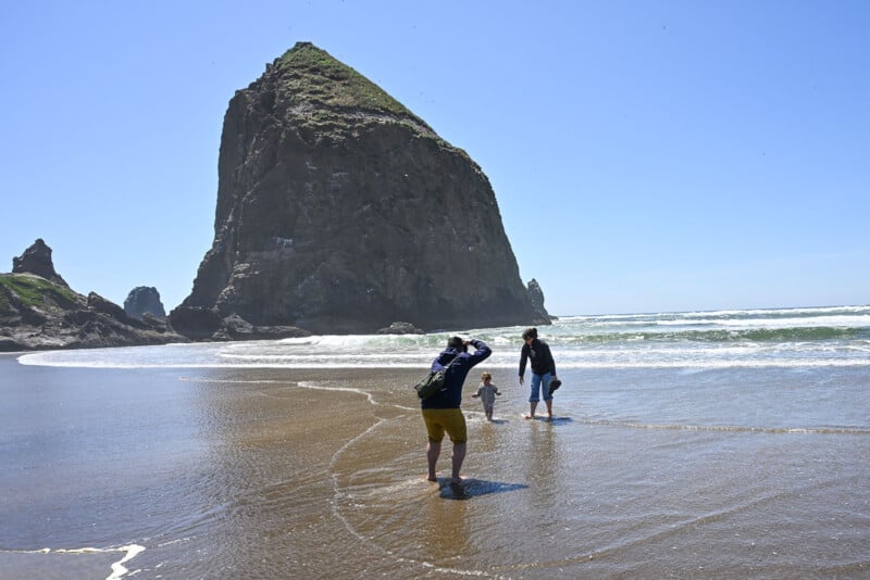 A man takes a photo of two people, one child and one adult, wading in shallow water near a large rock formation on a sunny beach. Waves crash in the background under a clear blue sky.