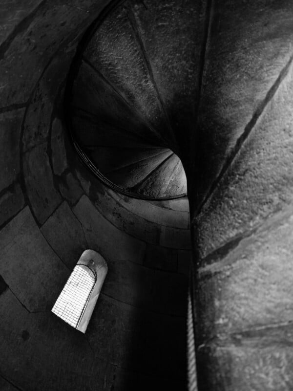 Black and white image of a spiral stone staircase viewed from a narrow-angle. Light illuminates through a small rectangular window with a metal grid, highlighting the textured stone steps and walls, creating an intriguing pattern of light and shadow.