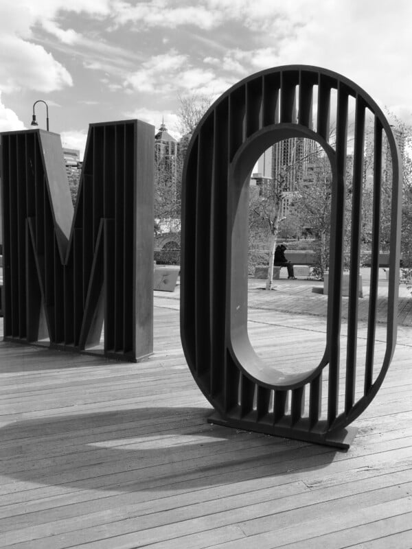 Large metal letters "M" and "O" stand upright on a wooden deck in an outdoor urban park, with trees and tall buildings visible in the background. The image is in black and white.