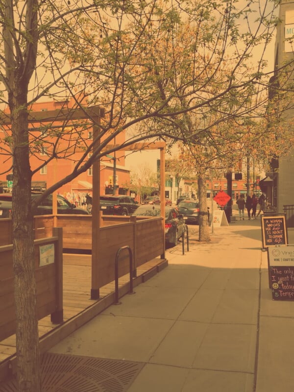 A sidewalk lined with trees and a wooden patio on the left, with people walking in the background on a sunny day. Cars are parked along the street and a chalkboard menu stands near a building on the right.