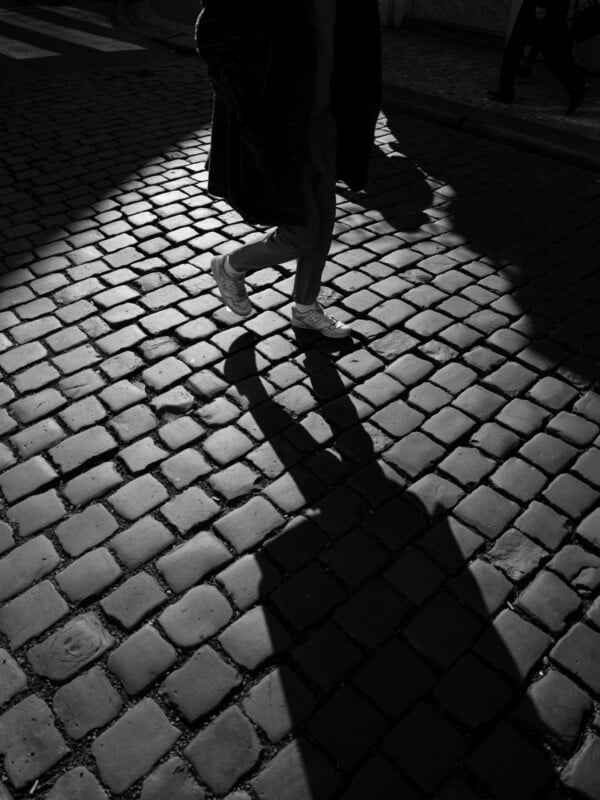 Black and white image of a person walking on a cobblestone street, casting a long shadow. The person is wearing a coat and sneakers, and the street is partly illuminated, creating contrast with darker areas.