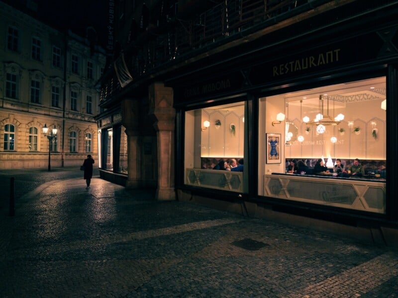 A dimly lit street scene captures a person walking past a warmly illuminated restaurant with large windows. Inside, diners sit at tables beneath modern light fixtures. The surrounding buildings exhibit classic architecture.