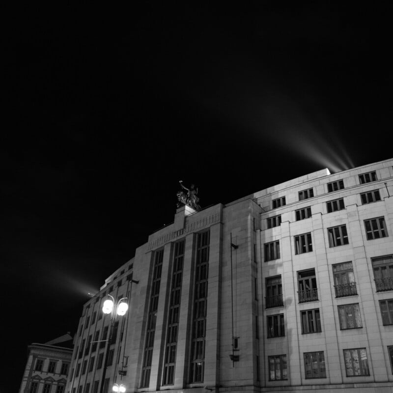 Black and white photo of a tall, historical building with ornate details at night. Bright lights illuminate the upper facade and a statue on the roof. Streetlights are visible in the foreground.