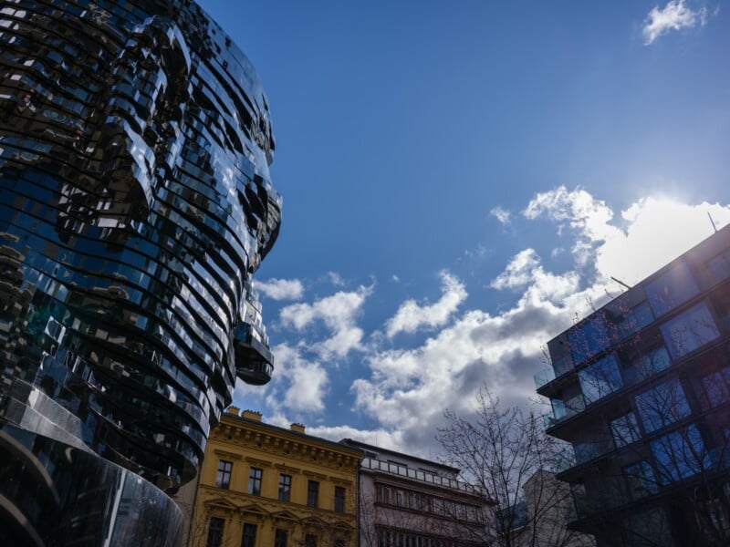 The image shows a layered, mirrored sculpture of a face against a blue sky with clouds and sunlight. Surrounding buildings include a yellow one with ornate architecture. Reflective glass panels of a modern building are also visible.