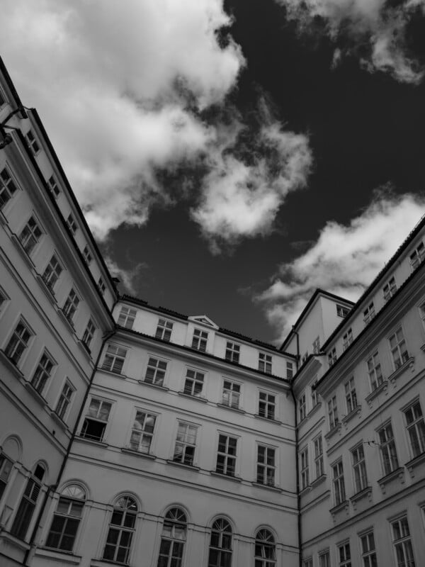 Black and white image of a historic building's courtyard with multiple windows. The view looks up toward the sky, showcasing dramatic clouds framed by the building's walls.