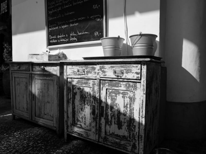 A black and white photo of rustic cabinets set against a wall with a chalkboard menu above. Two buckets sit on top of the weathered surface. Sunlight casts shadows, highlighting the texture of the wood and the cobbled street below.