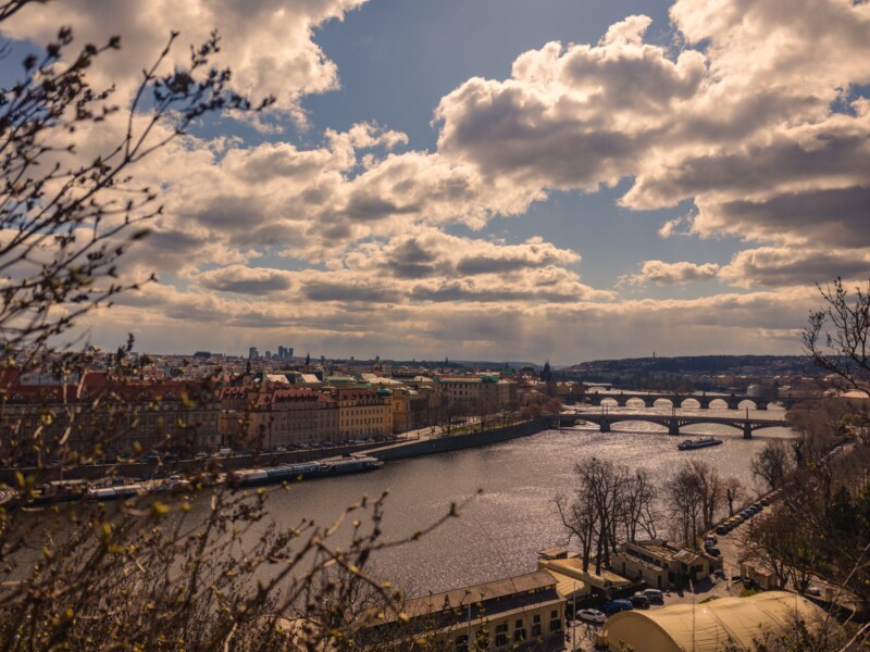 A scenic view of a city with multiple bridges spanning a river under a partly cloudy sky. On the left, historic buildings line the riverbank, and leafless trees frame the foreground. The sun casts a warm glow over the landscape.