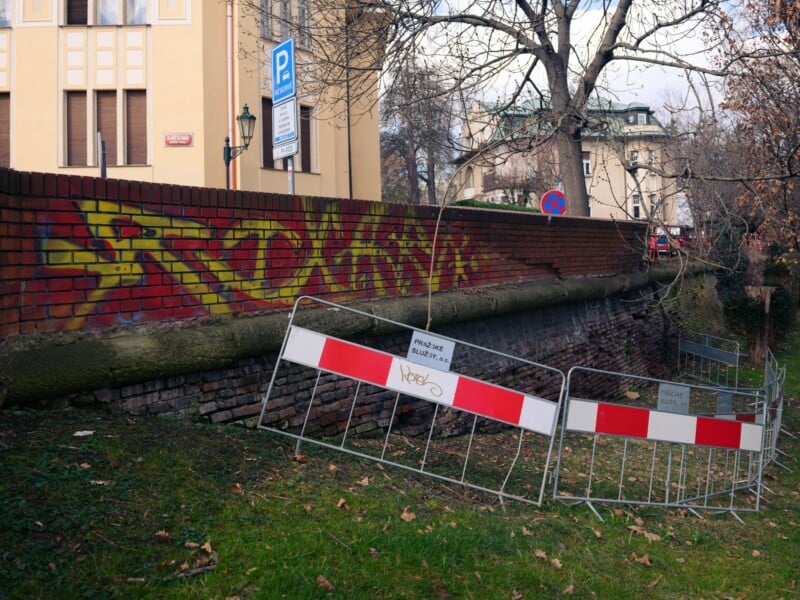 Brick wall with yellow and red graffiti next to a sidewalk. A metal barrier with red and white stripes blocks access to the grassy area beside the wall. Nearby, a parking sign and traffic sign are visible. Bare trees are in the background.