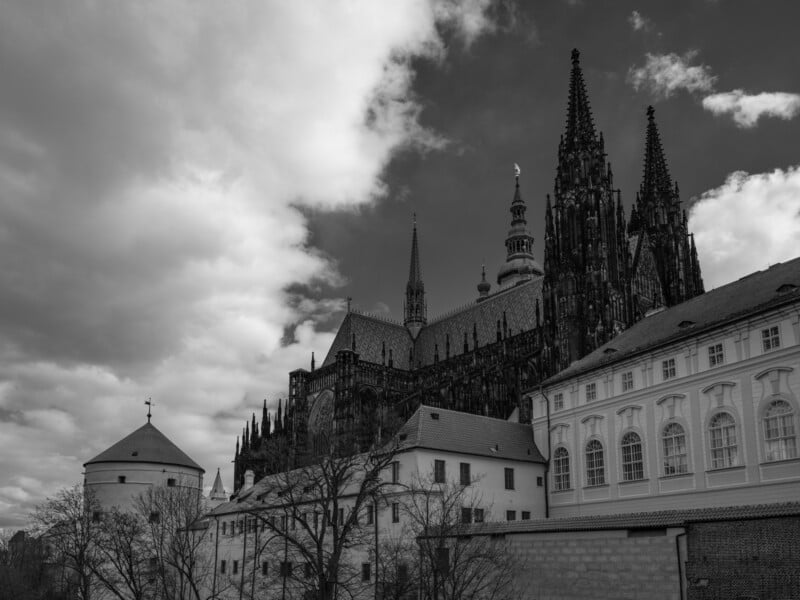 Black and white photo of a Gothic cathedral with tall spires on a cloudy day. Foreground includes old buildings and leafless trees. The dramatic sky adds a moody atmosphere to the scene.