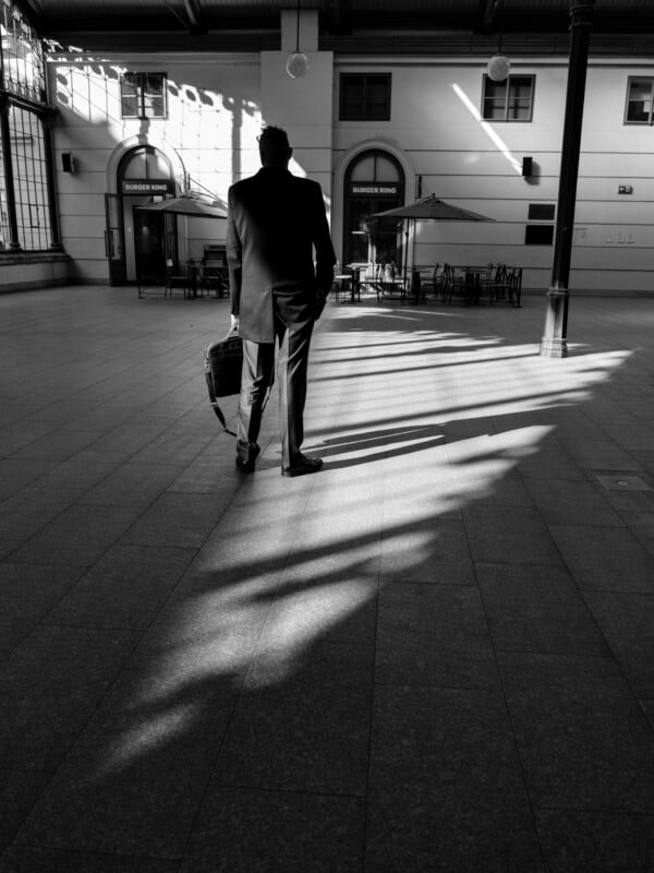 A person in a suit stands alone in a spacious, sunlit corridor with a briefcase. Long shadows stretch across the floor from large arched windows, creating a dramatic effect.