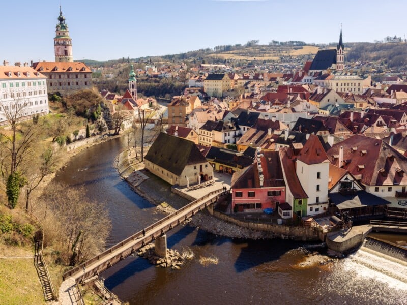 Aerial view of Český Krumlov, Czech Republic, showcasing the Vltava River, a wooden bridge, and the town's historic architecture with colorful buildings and church spires under a clear blue sky.