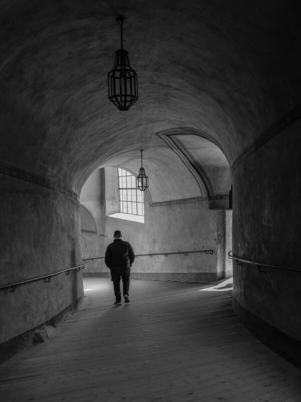 A person walks down a dimly lit, arched corridor with textured walls. Two hanging lanterns illuminate the space. Sunlight streams through a large window at the end of the corridor, casting light on the floor. The image is in black and white.