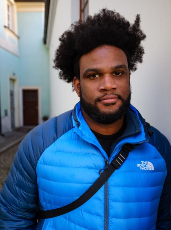 A person with natural curly hair and a beard is wearing a bright blue puffer jacket with a black shoulder strap, standing in a narrow outdoor alleyway. The background features light-colored buildings with a wooden door.