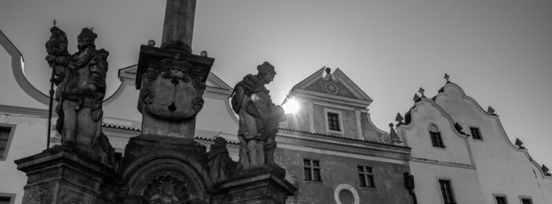 Black and white image of a historic building with ornate architecture and statues. The statues stand beside a column in the foreground. The sun peeks through, casting shadows across the scene, enhancing the dramatic contrast.