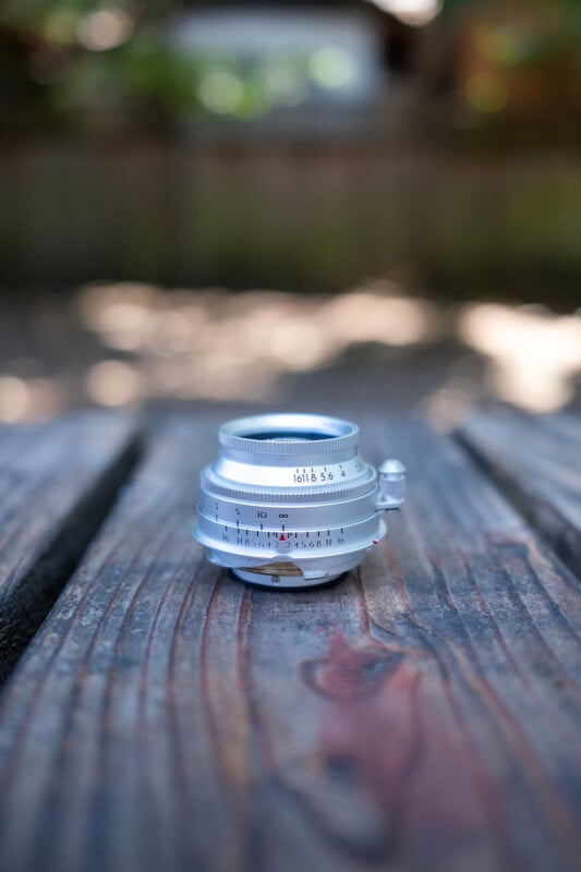 A silver camera lens sits upright on a weathered wooden surface outdoors, with a blurred background of greenery and a fence.