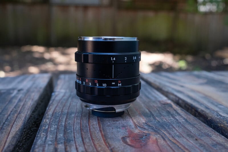 A camera lens with focus and aperture markings sits upright on a weathered wooden table outdoors, with a blurred fence and greenery in the background.