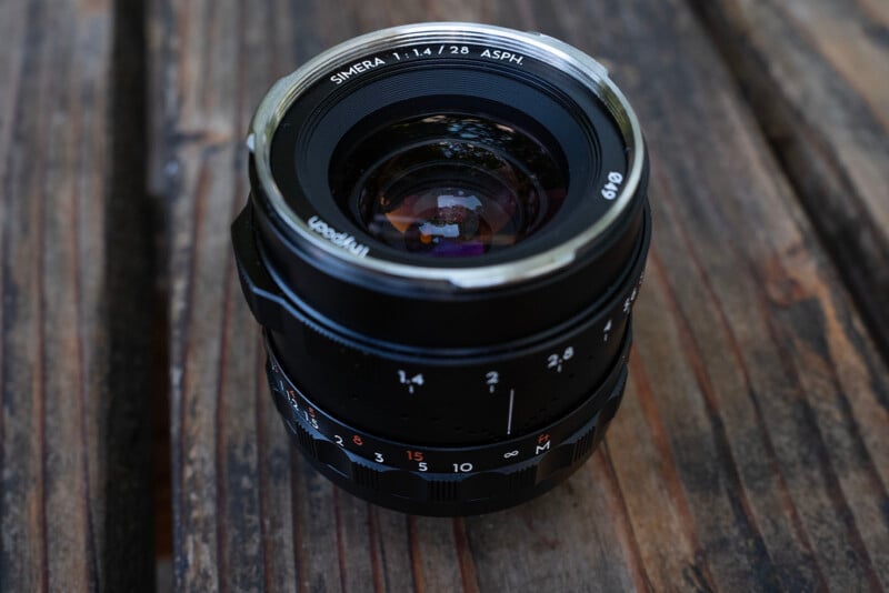 A close-up photo of a Smera 28mm f/1.4 camera lens placed on a weathered wooden surface, showing aperture and focus markings clearly on the barrel.