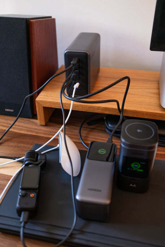 A desk with various electronic devices and chargers plugged in, including power banks, a computer mouse, and cables connected to a charging hub on a wooden shelf.