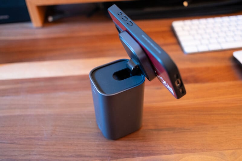 A black smartphone is held upright in a rectangular, black charging stand on a wooden desk, with a white keyboard blurred in the background.
