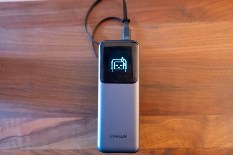 A UGREEN portable power bank sits on a wooden surface, connected by a black cable. Its display screen shows a battery icon, indicating it is charging.