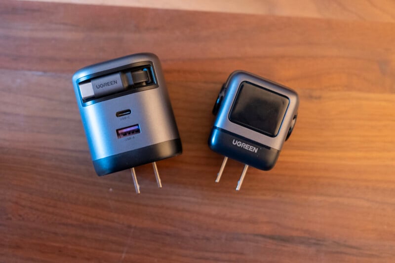 Two UGREEN travel adapters with retractable plugs are placed on a wooden surface. The adapter on the left shows USB ports, while the one on the right displays the brand logo and has its plug extended.