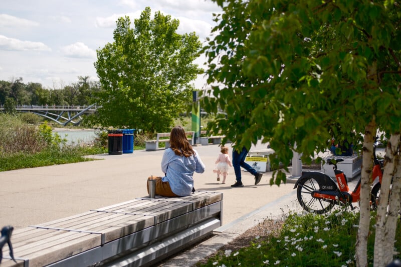 A woman sits on a wooden bench near trees and flowers in a park. A man walks ahead with a small child, and a red rented bicycle is parked nearby. A bridge and river are visible in the background.