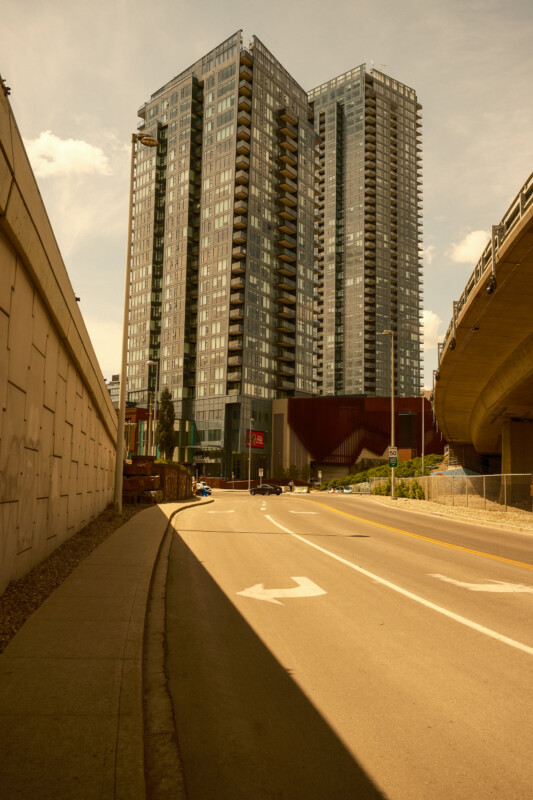 A tall, modern glass apartment building stands between a highway overpass and a concrete wall on a sunny day, with a curved road and arrow markings in the foreground.