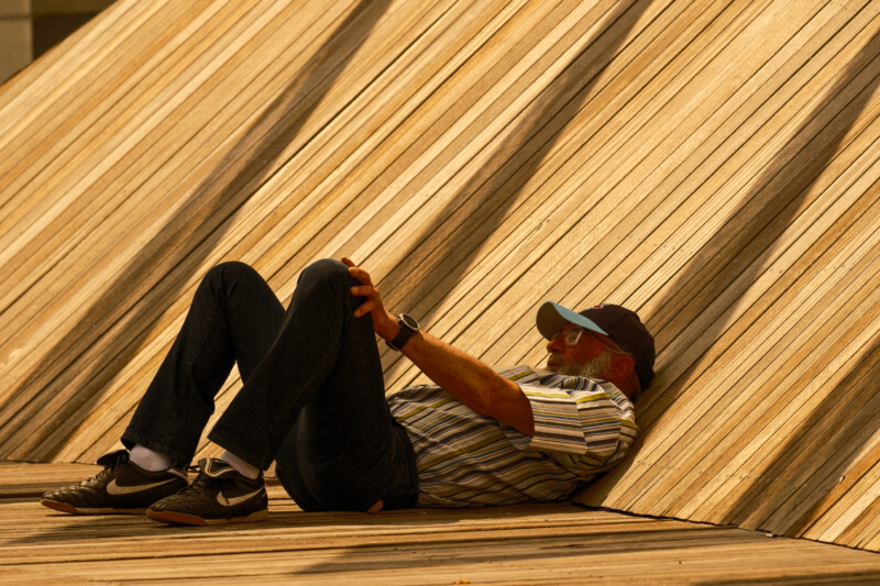 A man wearing a cap, striped shirt, and sneakers reclines on his back against a wooden slatted structure, knees bent, arms resting on his legs, in warm sunlight.