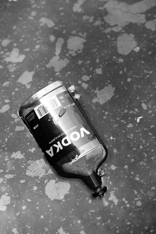A black and white photo of an empty vodka bottle lying on a wet, textured surface with scattered water spots and stains.