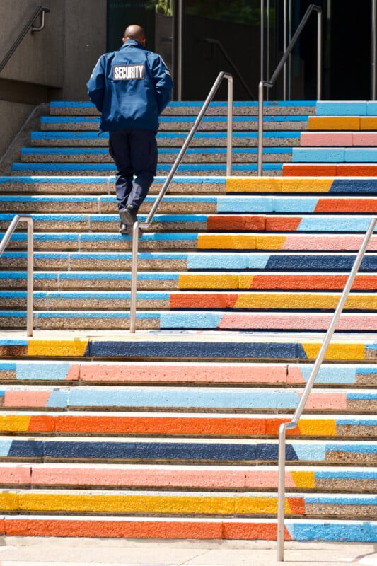A security guard walks up a set of outdoor stairs painted with bright, colorful stripes and geometric patterns in blue, orange, yellow, and red. Metal railings line both sides of the stairs.