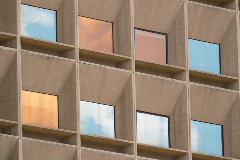 Close-up of a grid of concrete-framed windows on a building facade, with some window panes reflecting blue sky and clouds, and others reflecting a warm orange hue.