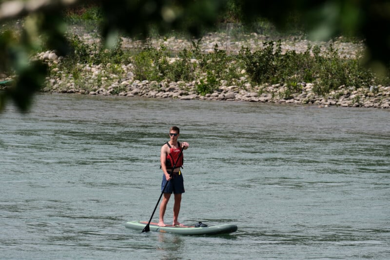 A person wearing sunglasses and a red life vest stands on a paddleboard in a river, holding a paddle, with a rocky shoreline and greenery in the background.