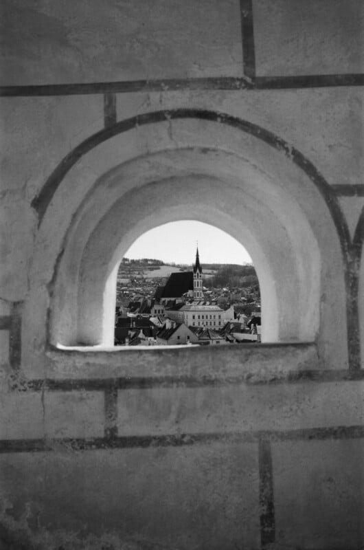 A black and white photo of a town seen through an arched stone window, with a prominent church and steeple in the center and rooftops spread out below under a clear sky.