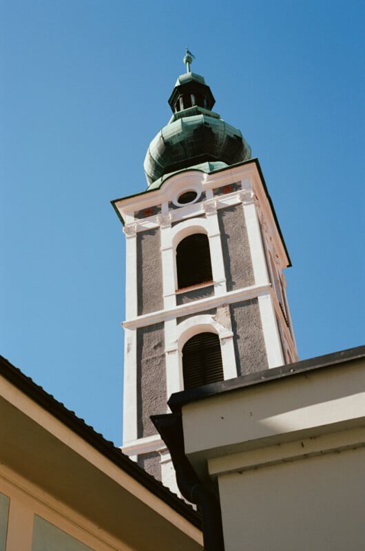 A tall church tower with arched windows and a green dome rises against a clear blue sky, partially framed by the edges of nearby rooftops.