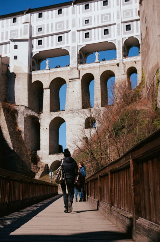 A person walks on a wooden bridge toward a tall, multi-level stone and white building with large arches and windows, with a clear blue sky above. Other people are visible in the background.