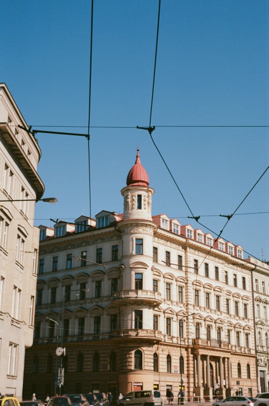 A historic, ornate building with a red conical tower roof stands under a clear blue sky, framed by tram wires overhead and surrounded by other urban buildings.