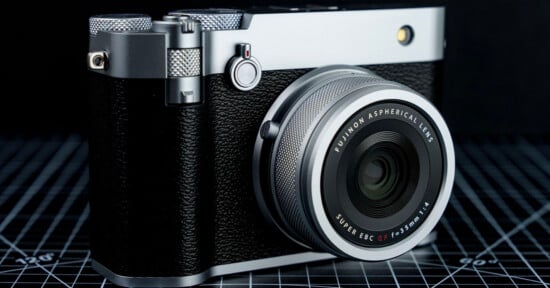 A silver and black vintage-style camera is placed on a grid-patterned surface. The camera features a large lens with markings "Fujinon Aspherical Lens" and "Super EBC 23mm f/2." The body has textured grips and metal dials.