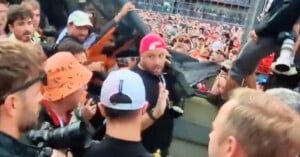 A large crowd of people, some wearing hats and holding cameras, gather around a barricade at an outdoor event. A man in a red cap stands at the center, facing the camera, while photographers capture the scene.