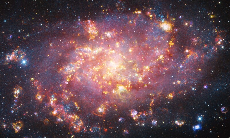 A bright, colorful spiral galaxy with glowing clouds of gas and dust, sparkling stars, and clusters scattered throughout the image against the dark backdrop of space.