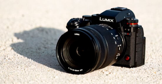 A Panasonic Lumix camera with a large lens sits on sandy ground. The camera is black with red accents and has "S1R" written on the grip area. The lens cap displays the Lumix logo, and the scene is lit by sunlight.