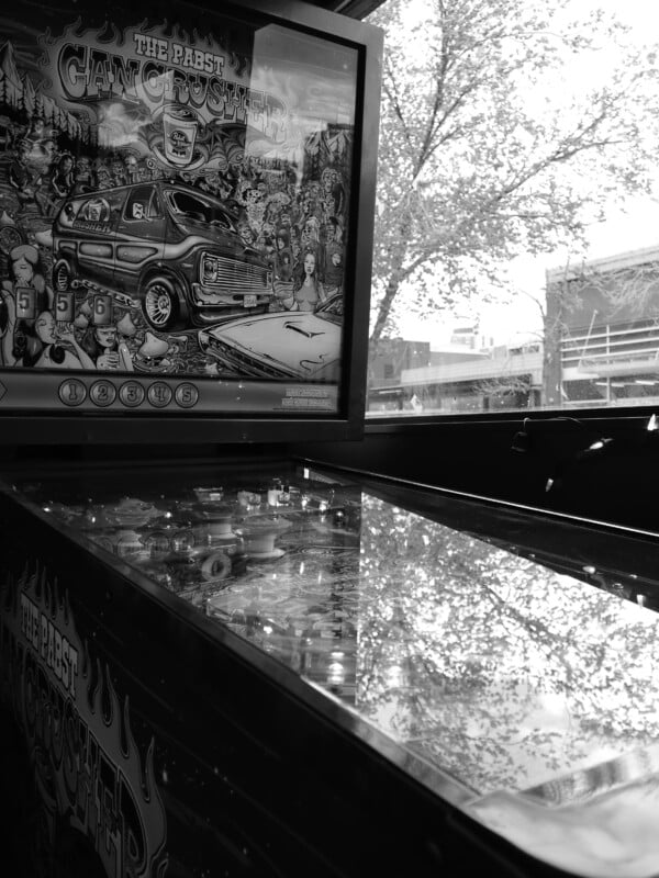 Black and white photo of a pinball machine titled “The Pabst Giant Crusher,” with its glass surface reflecting trees and a building visible through a nearby window.
