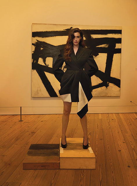 A woman in a black and white asymmetrical coat stands on stacked wooden blocks in an art gallery, with an abstract black and white painting on the wall behind her.