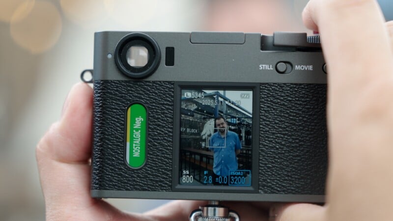 Close-up of a digital camera’s screen displaying a preview of a man in a blue shirt standing outdoors, with the camera settings and interface visible. A person’s hands are holding the camera.